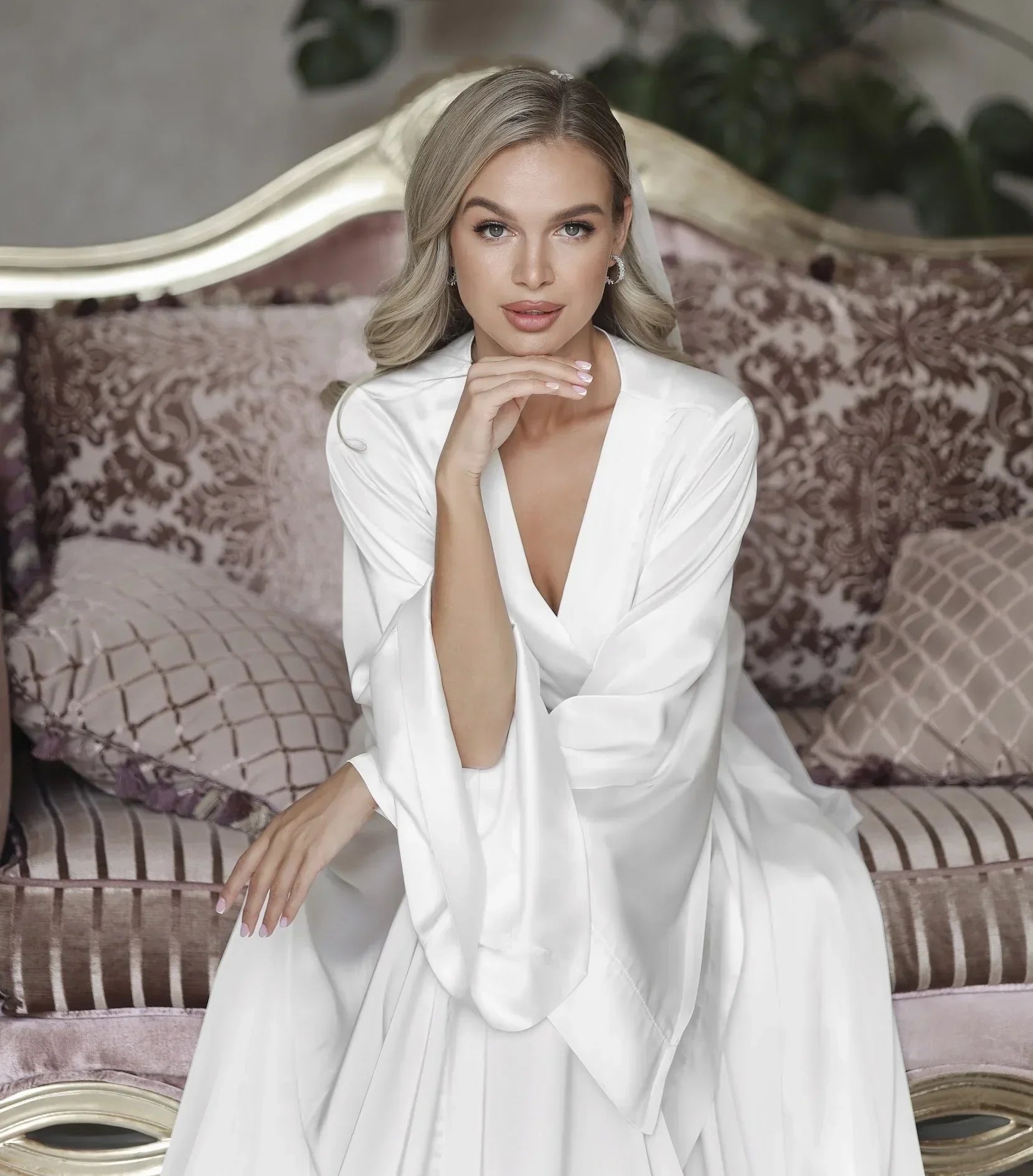 Bride in white silk bridal robe sitting on an elegant sofa, perfect for wedding morning.
